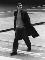 Placeholder for: Person in a long coat and sweater walking confidently on a city street, captured in black and white.