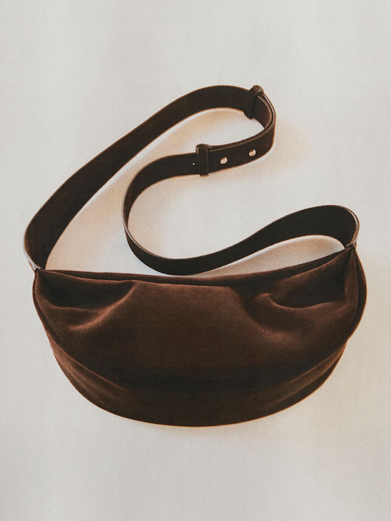 A brown velvet handbag with an adjustable leather shoulder strap and buckle, displayed on a light background.
