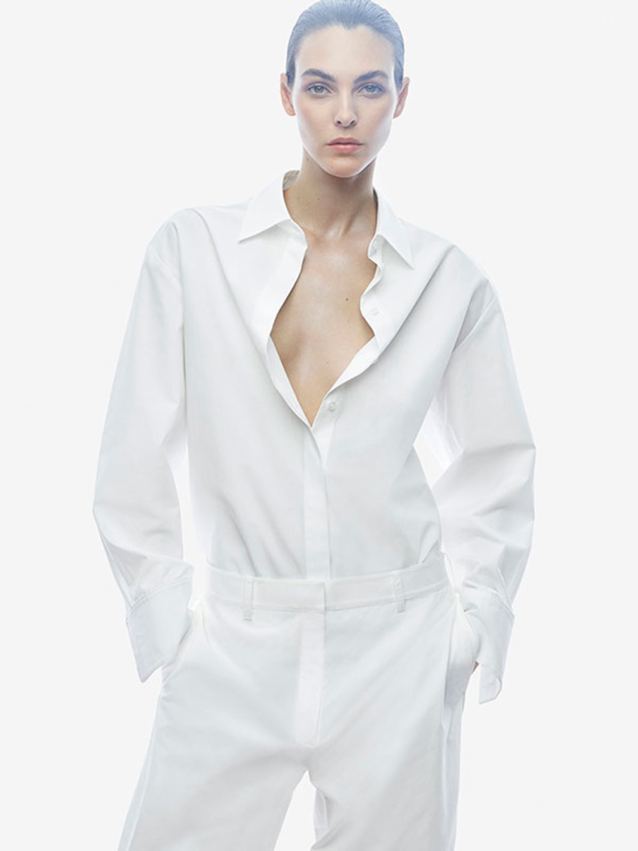 A person wearing a white shirt with an open collar and matching white pants, standing against a white background.