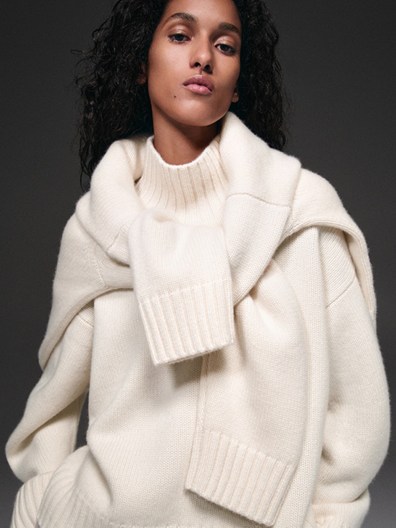 Person wearing a cream-colored, layered sweater, with relaxed curly hair, posing against a dark background.