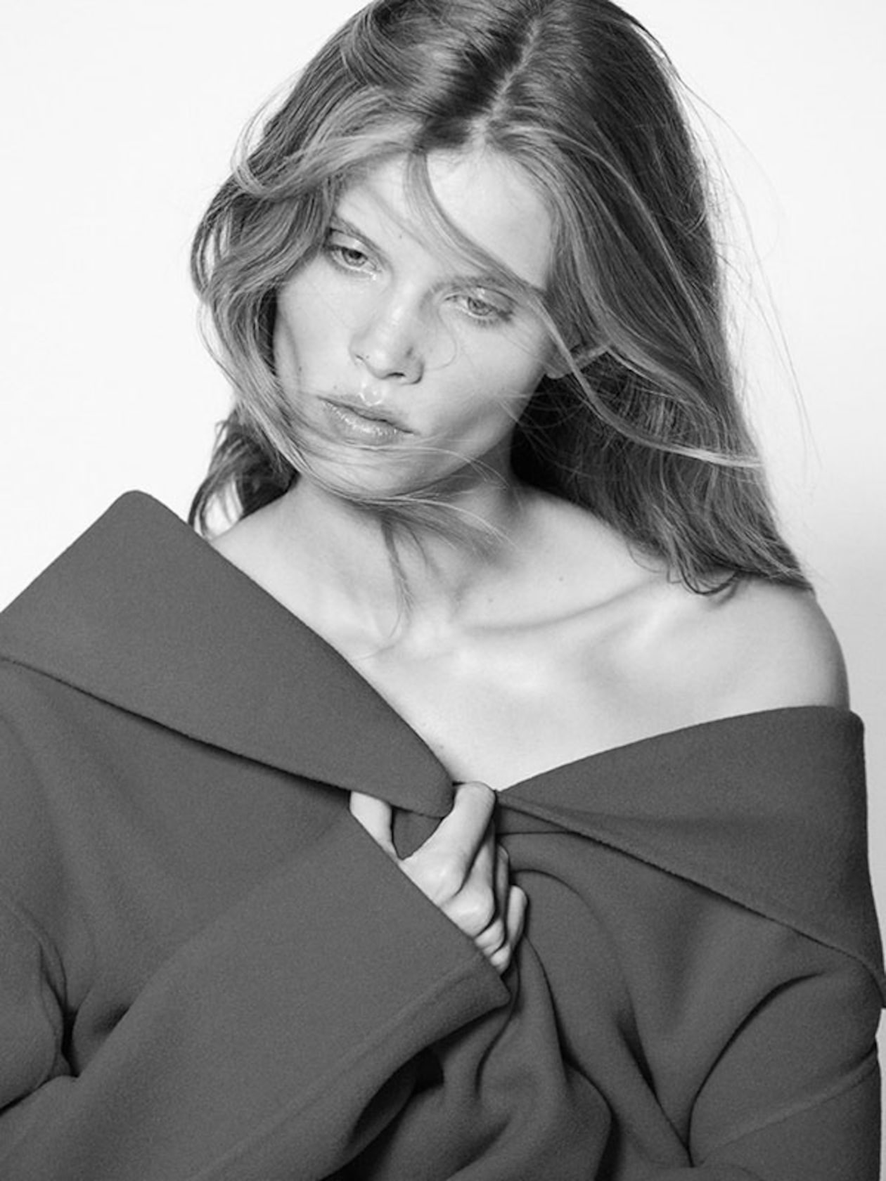 Black and white image of a woman with tousled hair, wearing an off-shoulder coat, looking down pensively.
