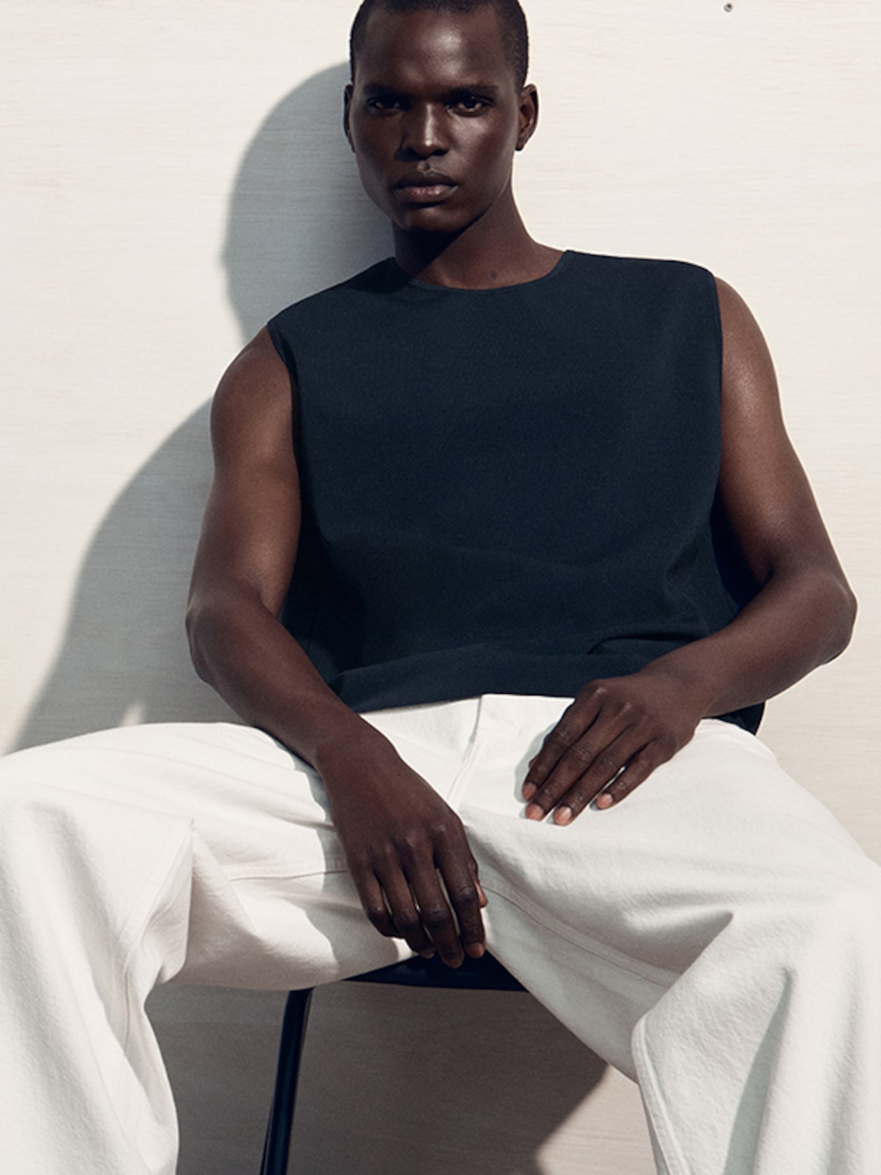 Person seated against a plain background, wearing a sleeveless black top and white pants, with a serious expression.
