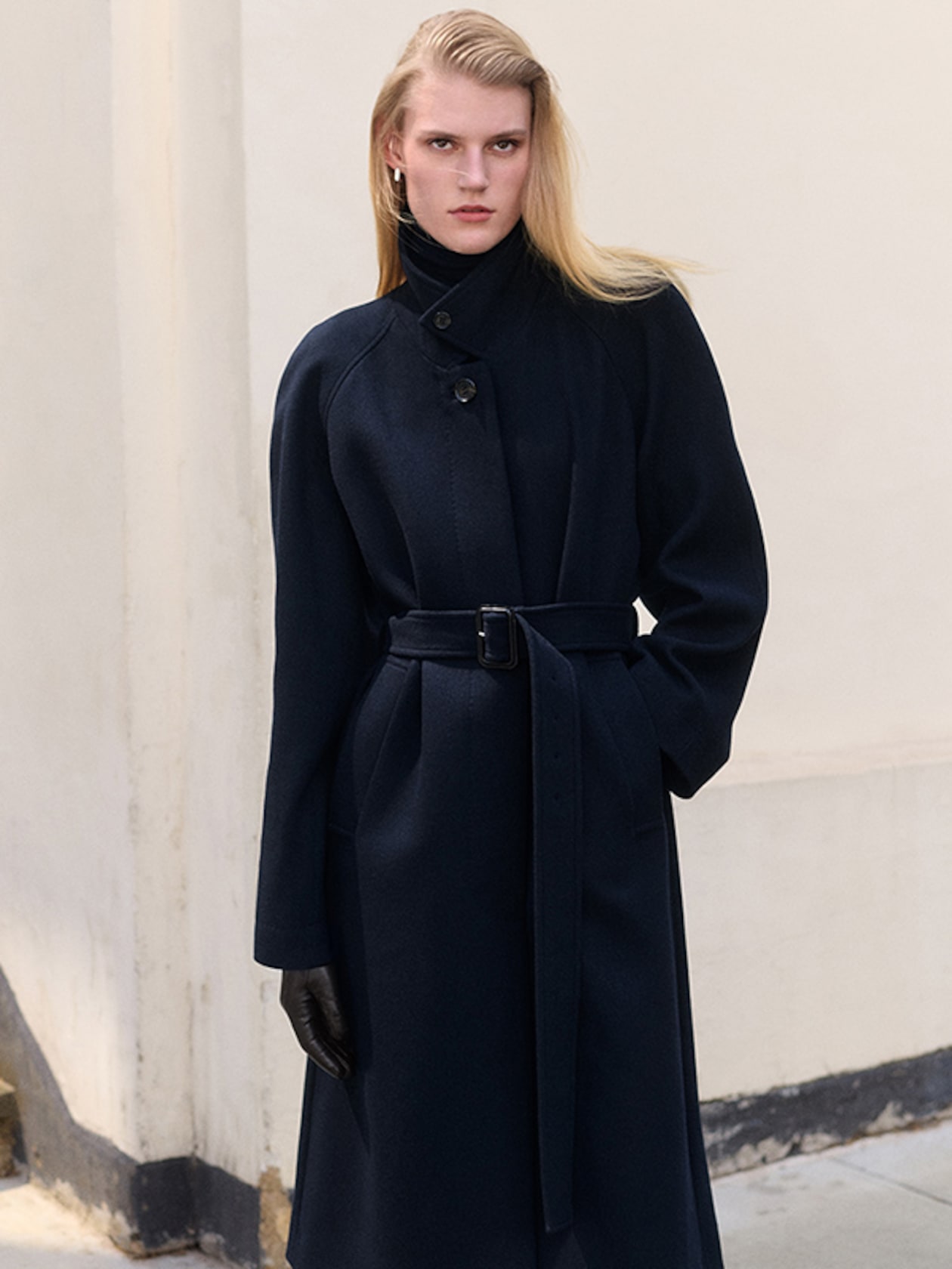 Person wearing a long, dark coat with a belt, black gloves, and light hair. They stand in front of a light-colored wall outdoors.
