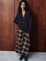 Placeholder for: Woman in a dark sweater and plaid skirt stands against a wooden wall, with curly hair and neutral expression, wearing ankle boots.