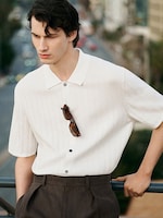 Placeholder for: A person with dark hair wearing a white collared shirt with sunglasses hanging, stands by a railing, gazing to the side.