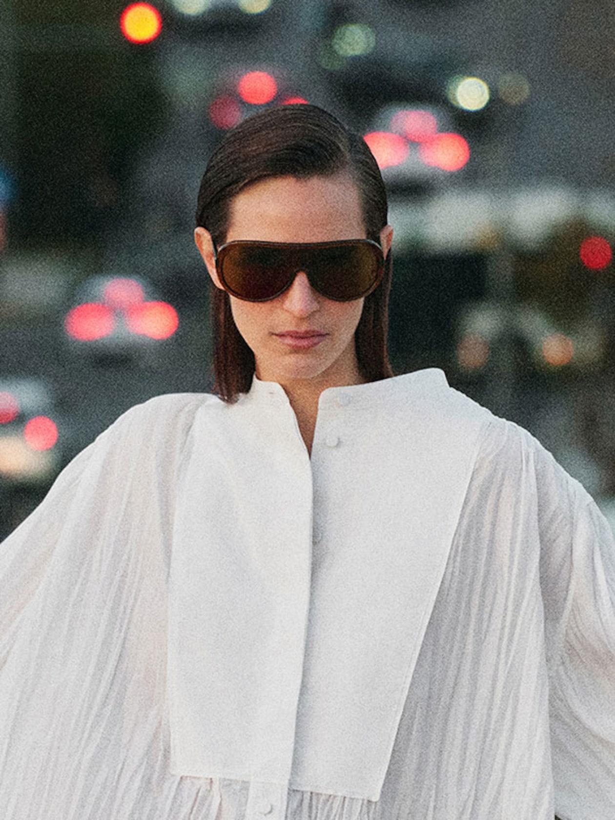A person wearing oversized sunglasses and a white pleated garment stands in front of a blurry urban background with bokeh lights.