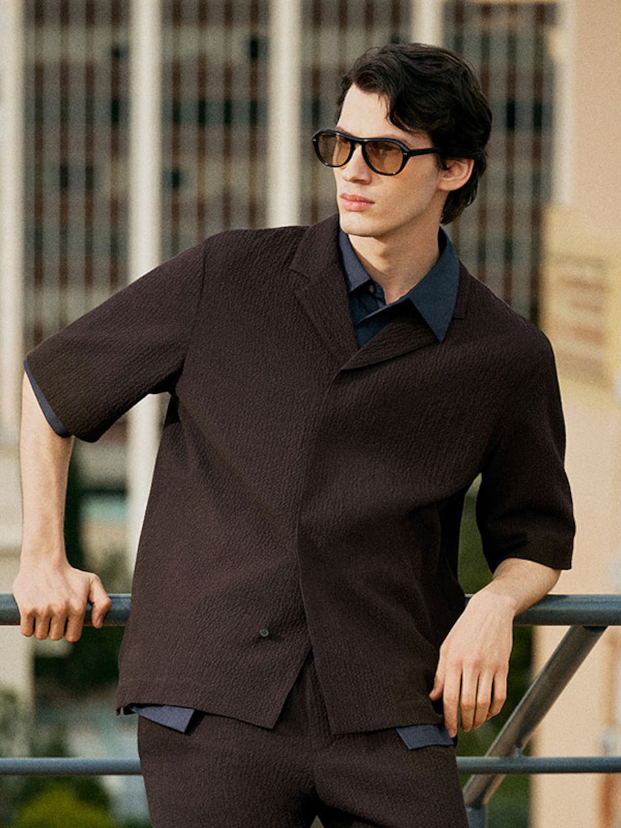 A man wearing sunglasses and a dark outfit leans against a railing outdoors, with a cityscape background.