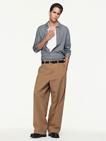 Placeholder for: Model in a checkered shirt over a white top, wearing beige wide-leg trousers and black shoes, standing with a casual pose.