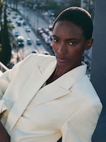 Placeholder for: Person wearing a light-colored suit, standing on a balcony with a blurred city street in the background.