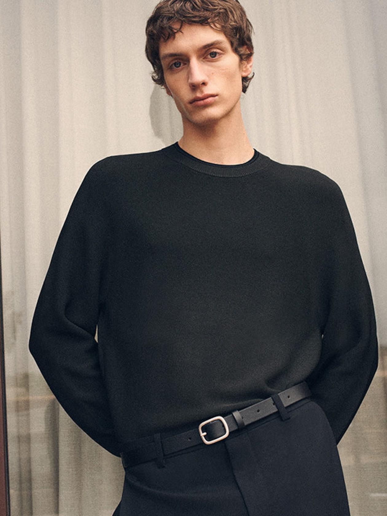 A person with short brown hair wearing a black sweater and black trousers stands indoors, hands in pockets, against a light curtain background.