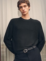 Placeholder for: A person with short brown hair wearing a black sweater and black trousers stands indoors, hands in pockets, against a light curtain background.