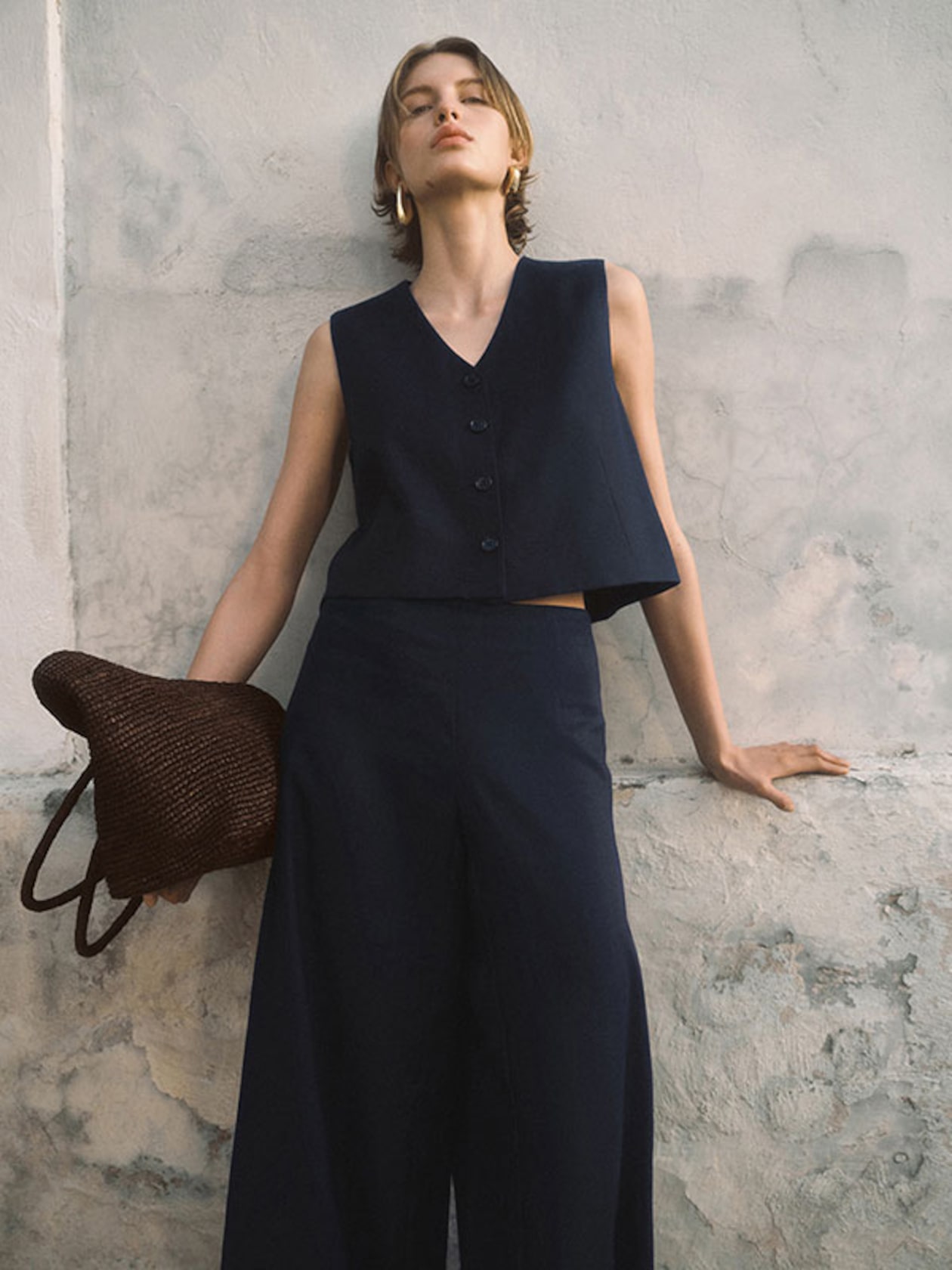 Person in a navy sleeveless top and wide-leg pants leans against a textured wall, holding a woven bag, with a relaxed expression.