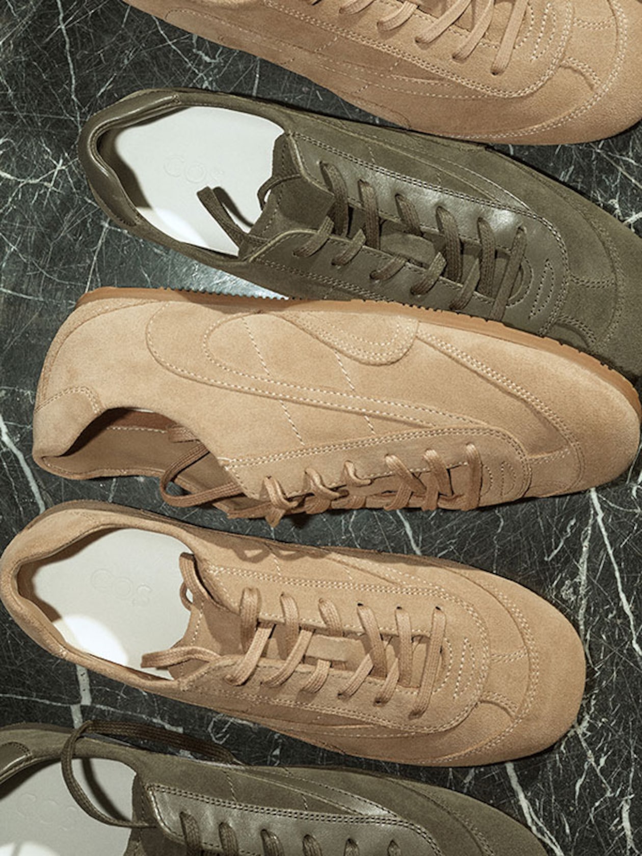 A top-down lifestyle shot of several pairs of COS low-top sneakers arranged on a dark green marbled surface. The collection includes muted sage green and warm taupe styles, highlighting a mix of textured suede and smooth leather panels with matching cotton laces.