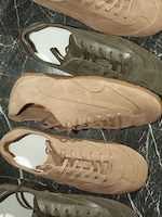 Placeholder for: A top-down lifestyle shot of several pairs of COS low-top sneakers arranged on a dark green marbled surface. The collection includes muted sage green and warm taupe styles, highlighting a mix of textured suede and smooth leather panels with matching cotton laces.