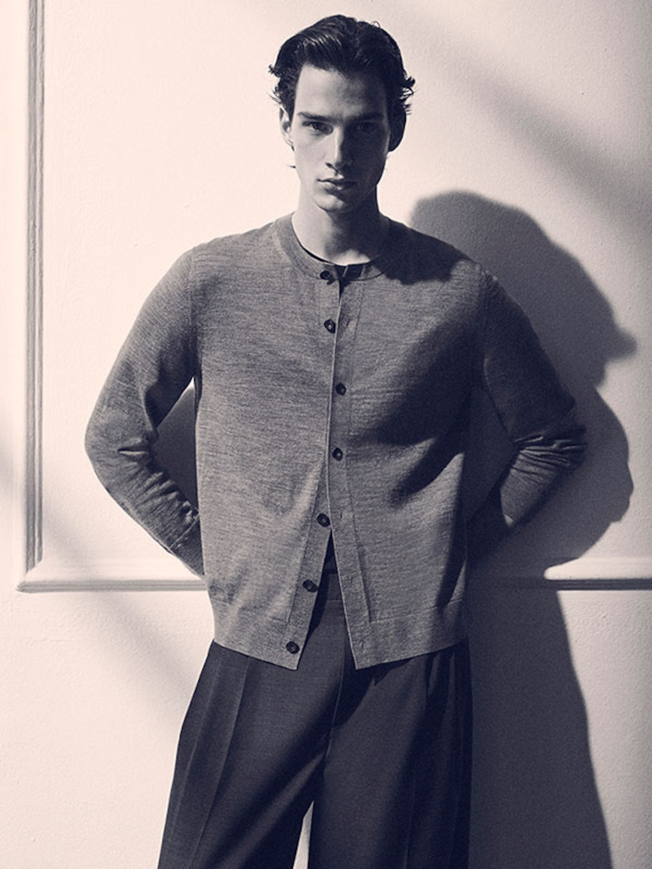 Man in a buttoned cardigan and trousers stands against a wall, casting a shadow. The image is in black and white.