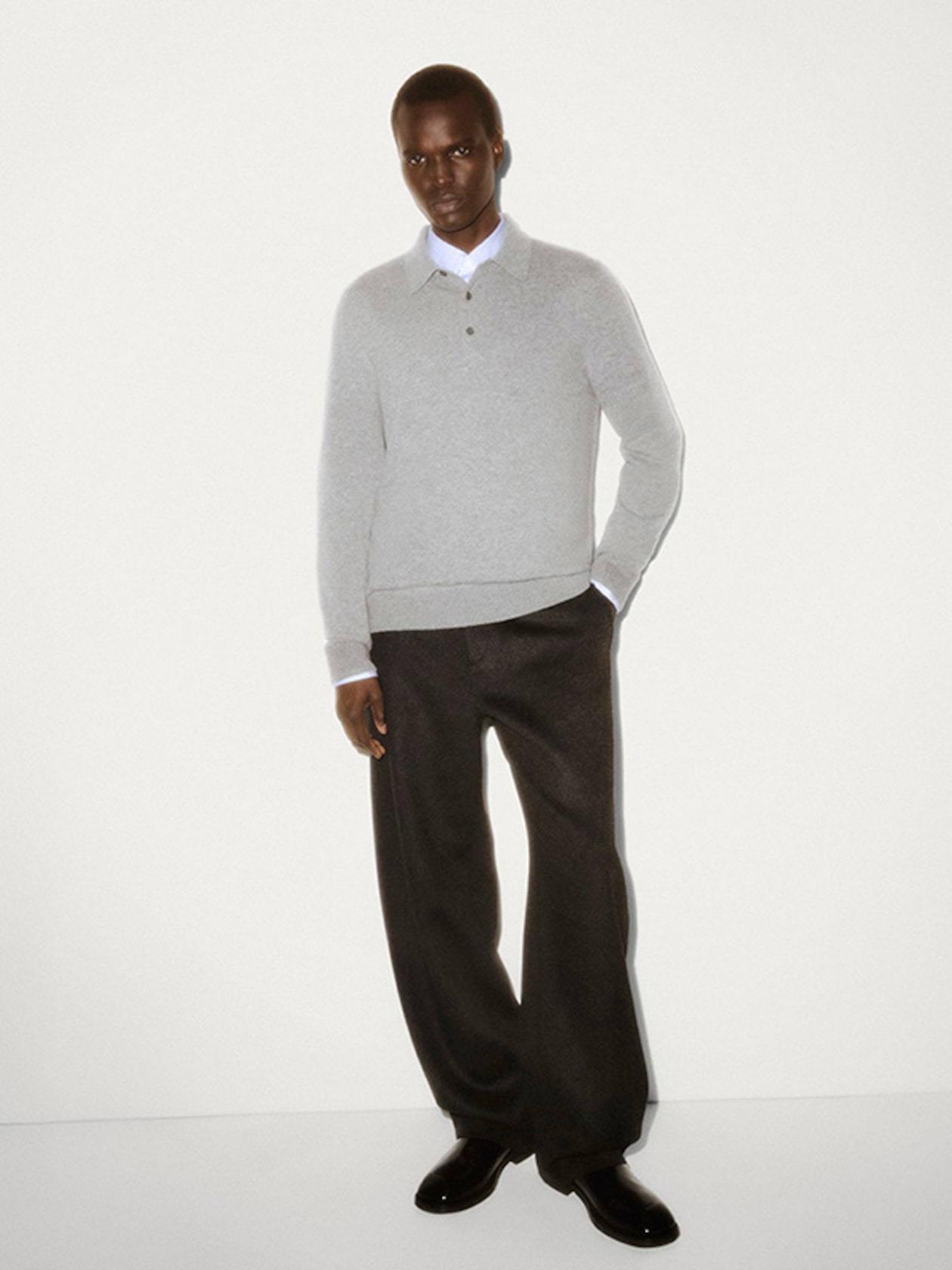 A person wearing a grey sweater, white shirt, and dark pants stands against a white background, hands in pockets, looking at the camera.