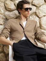 Placeholder for: A person wearing sunglasses, a beige jacket, and black pants sits in a white chair against a stone wall, exuding a relaxed, stylish vibe.