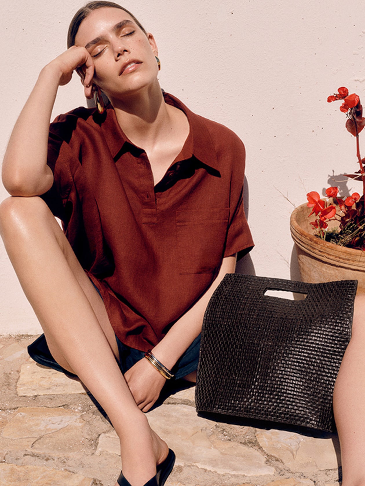 A sunlit profile shot of a woman with long wavy hair sitting outdoors against a white textured wall. She is wearing a bright red, knitted V-neck sleeveless cardigan vest with tonal buttons and black drawstring trousers. The look is accessorized with chunky silver hoop earrings and a thick silver bangle.