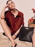 Placeholder for: A sunlit profile shot of a woman with long wavy hair sitting outdoors against a white textured wall. She is wearing a bright red, knitted V-neck sleeveless cardigan vest with tonal buttons and black drawstring trousers. The look is accessorized with chunky silver hoop earrings and a thick silver bangle.