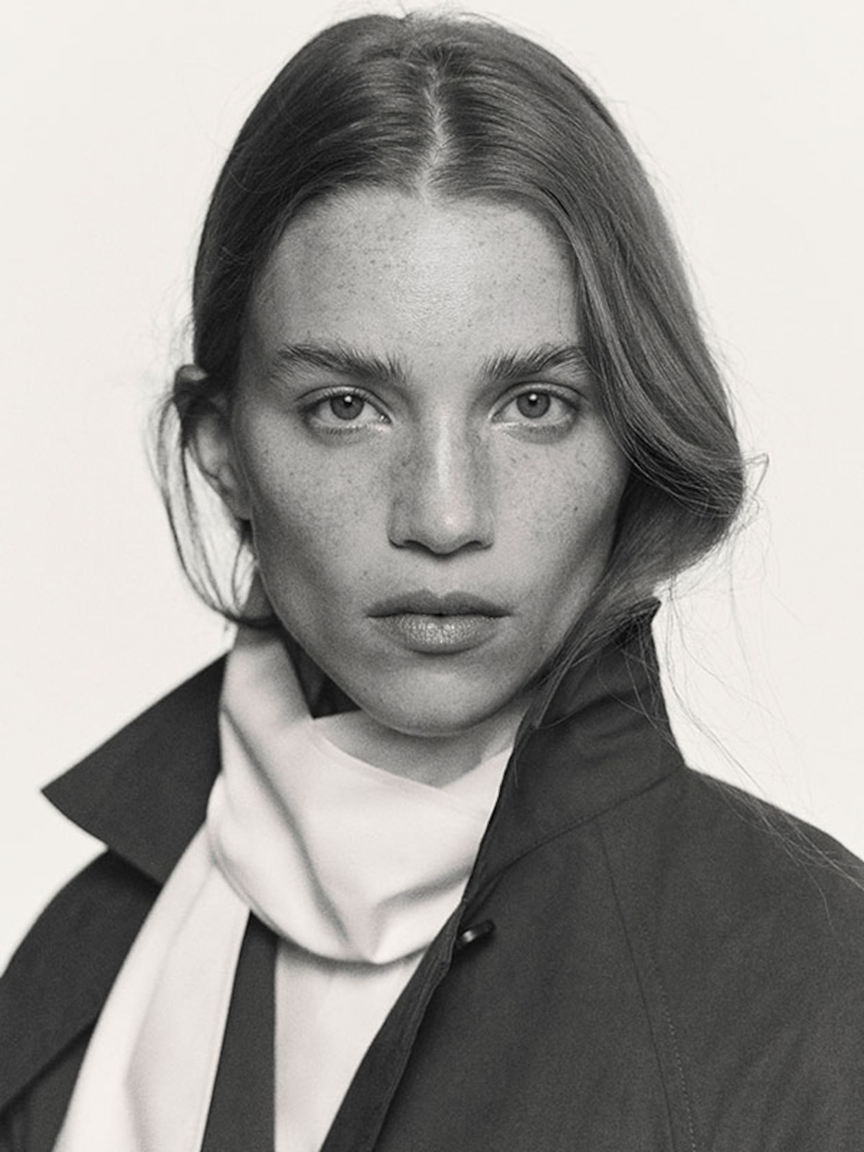 Black and white portrait of a woman with long hair, wearing a light scarf and dark coat, gazing directly at the camera.