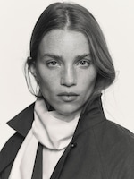 Placeholder for: Black and white portrait of a woman with long hair, wearing a light scarf and dark coat, gazing directly at the camera.