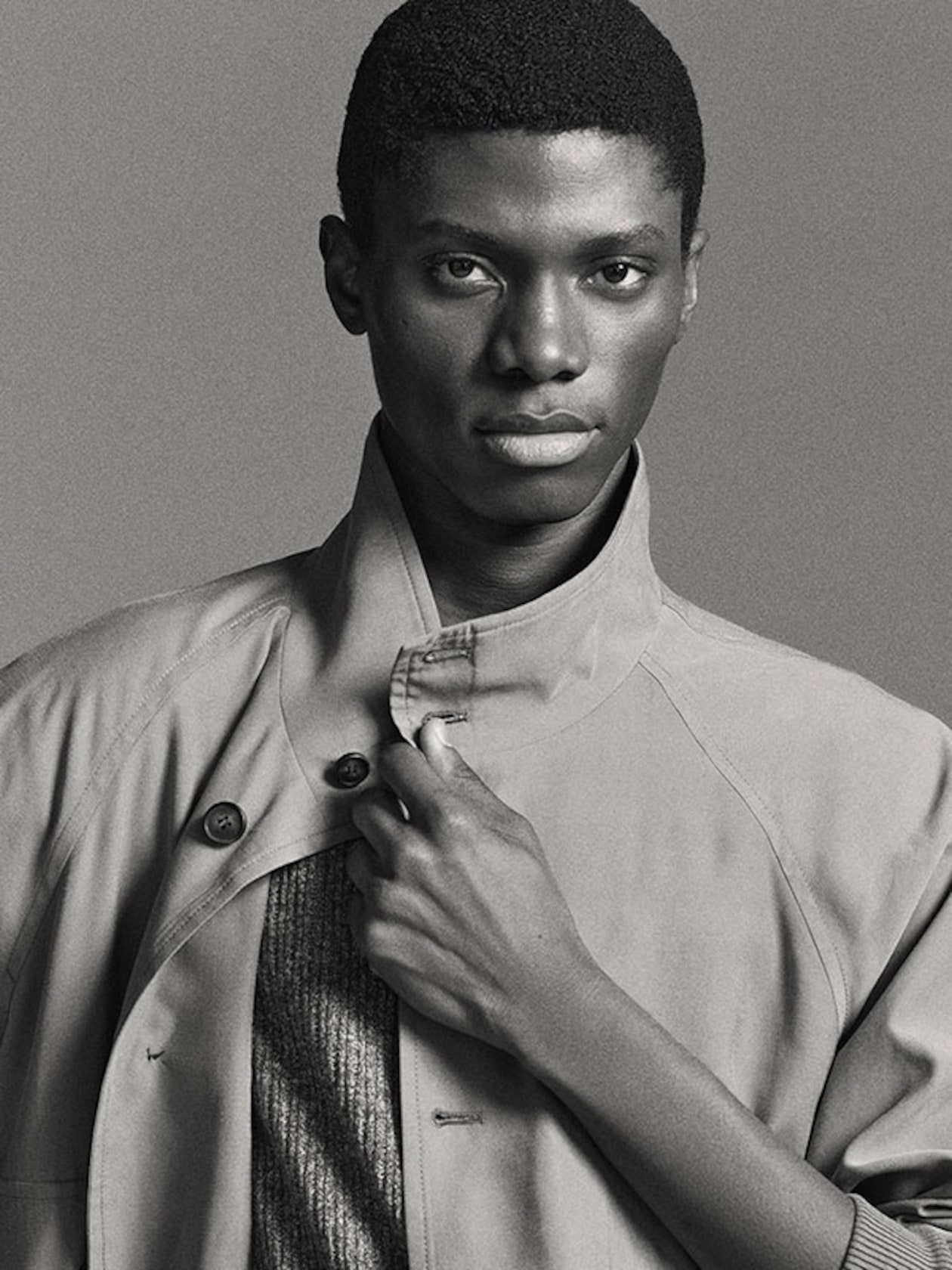 A person in a high-collared coat looks confidently at the camera, holding the collar with one hand in a black-and-white portrait.