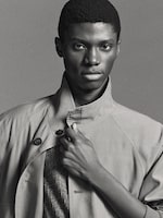 Placeholder for: A person in a high-collared coat looks confidently at the camera, holding the collar with one hand in a black-and-white portrait.
