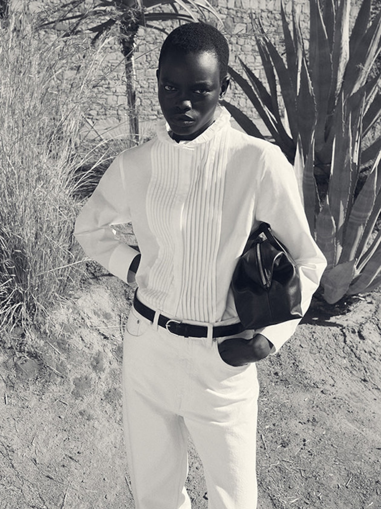 Person in a white outfit stands outdoors holding a bag, with a background of stone wall and large plants.