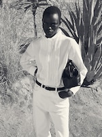 Placeholder for: Person in a white outfit stands outdoors holding a bag, with a background of stone wall and large plants.