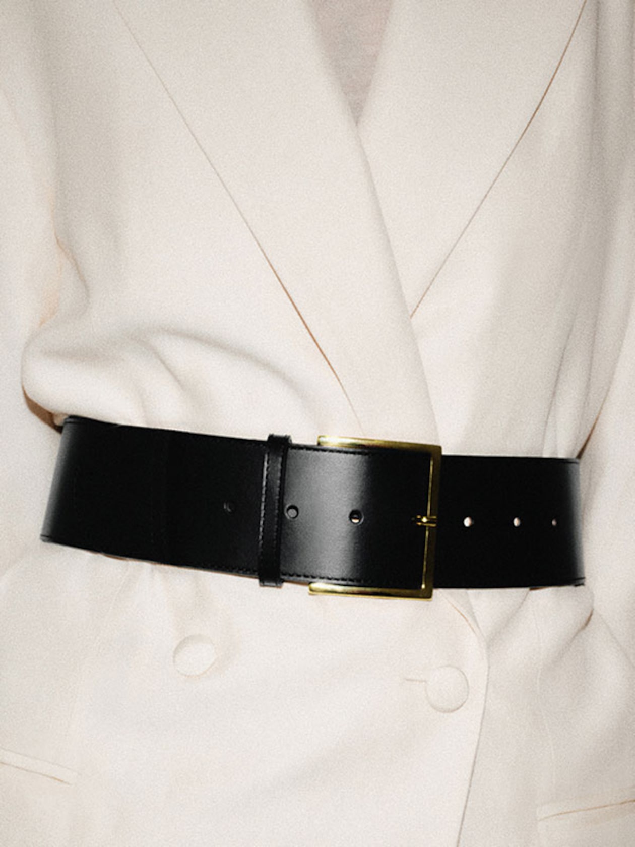 A black leather belt with a gold buckle cinches a white double-breasted coat, highlighting the tailored waistline.