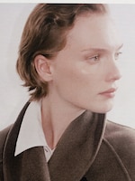 Placeholder for: Person with short hair wearing a white shirt and brown coat, gazing to the side with a neutral expression.