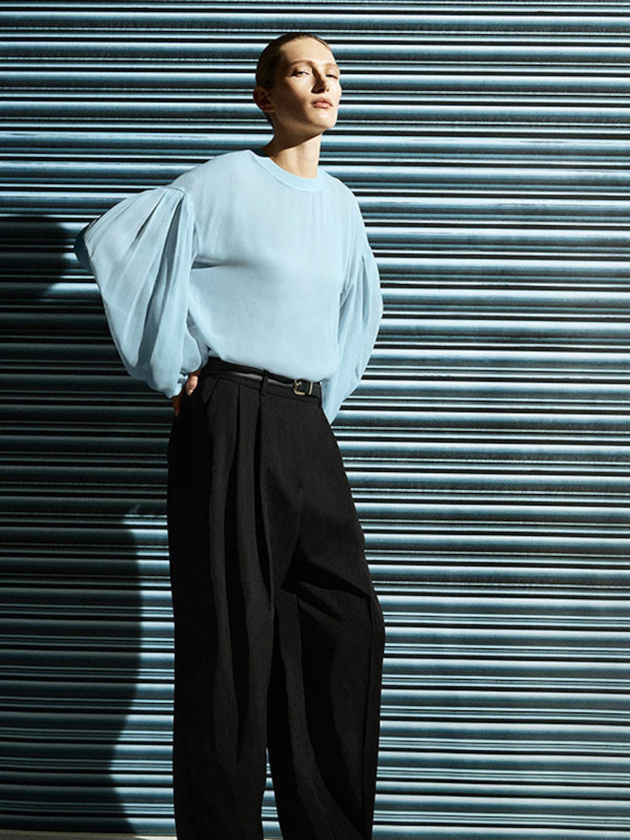Model in a light blue blouse with voluminous sleeves and black pants stands against a striped metal wall, looking to the side.