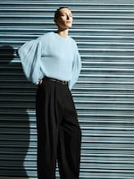 Placeholder for: Model in a light blue blouse with voluminous sleeves and black pants stands against a striped metal wall, looking to the side.
