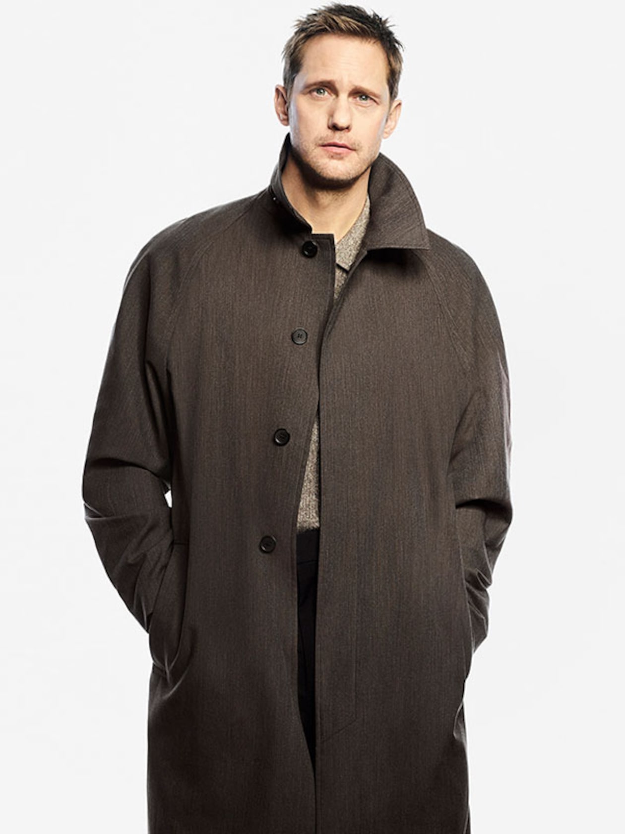 Man wearing a long buttoned black coat over a gray sweater, hands in pockets, standing against a plain light background.