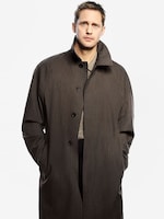 Placeholder for: Man wearing a long buttoned black coat over a gray sweater, hands in pockets, standing against a plain light background.