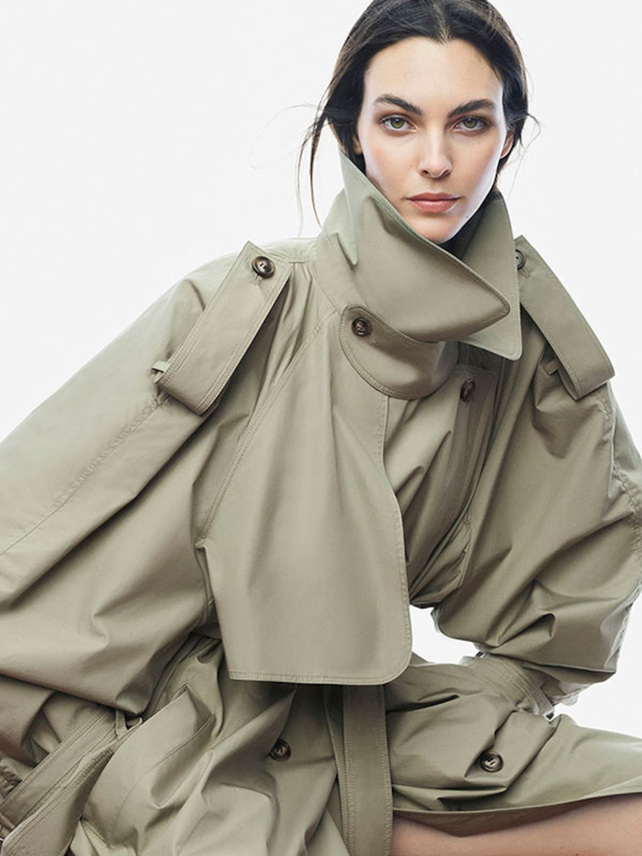 Woman in a tan oversized trench coat with an exaggerated collar and dramatic sleeves, looking directly at the camera against a plain background.