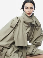 Placeholder for: Woman in a tan oversized trench coat with an exaggerated collar and dramatic sleeves, looking directly at the camera against a plain background.