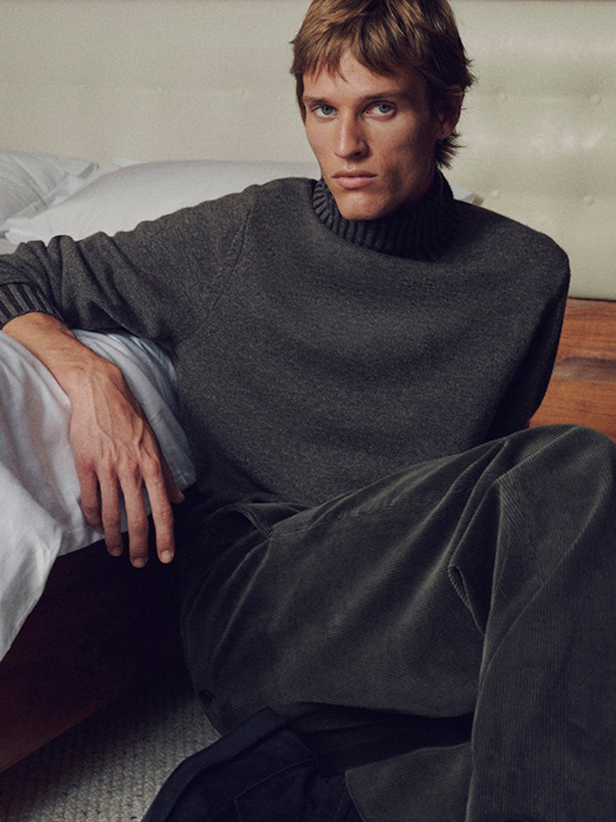 Person with short hair sitting on a bed, wearing a dark turtleneck sweater and corduroy pants, looking towards the camera.