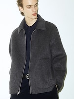 Placeholder for: Person wearing a dark gray herringbone jacket and a black shirt, looking to the side against a plain white background.