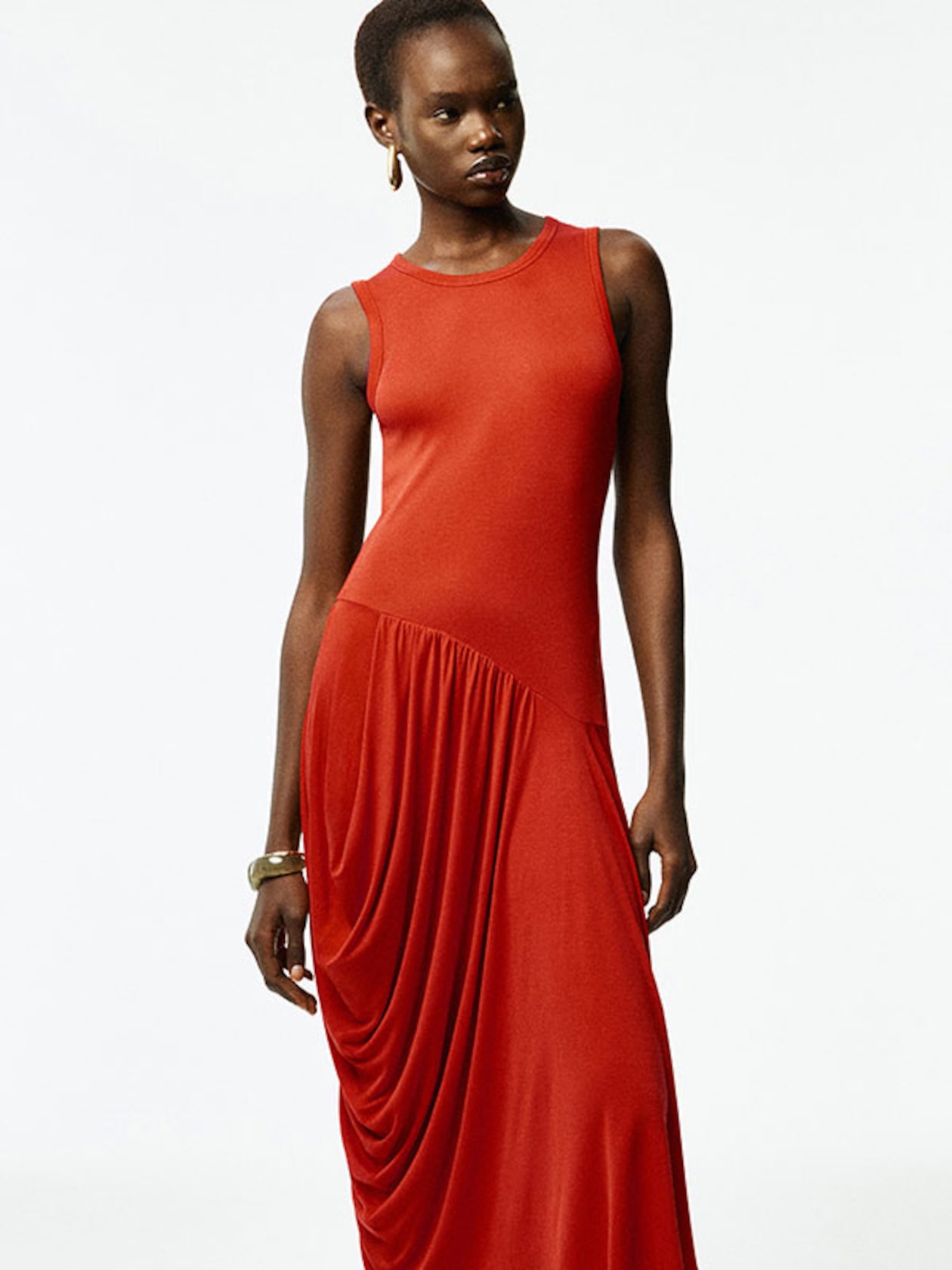 Person wearing a sleeveless, draped, long red dress with gold hoop earrings and a bracelet, standing against a plain white background.
