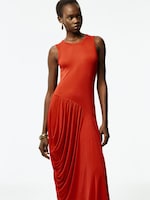 Placeholder for: Person wearing a sleeveless, draped, long red dress with gold hoop earrings and a bracelet, standing against a plain white background.