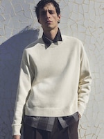 Placeholder for: Man in a cream sweater over a dark shirt stands against a textured wall, casting a shadow.