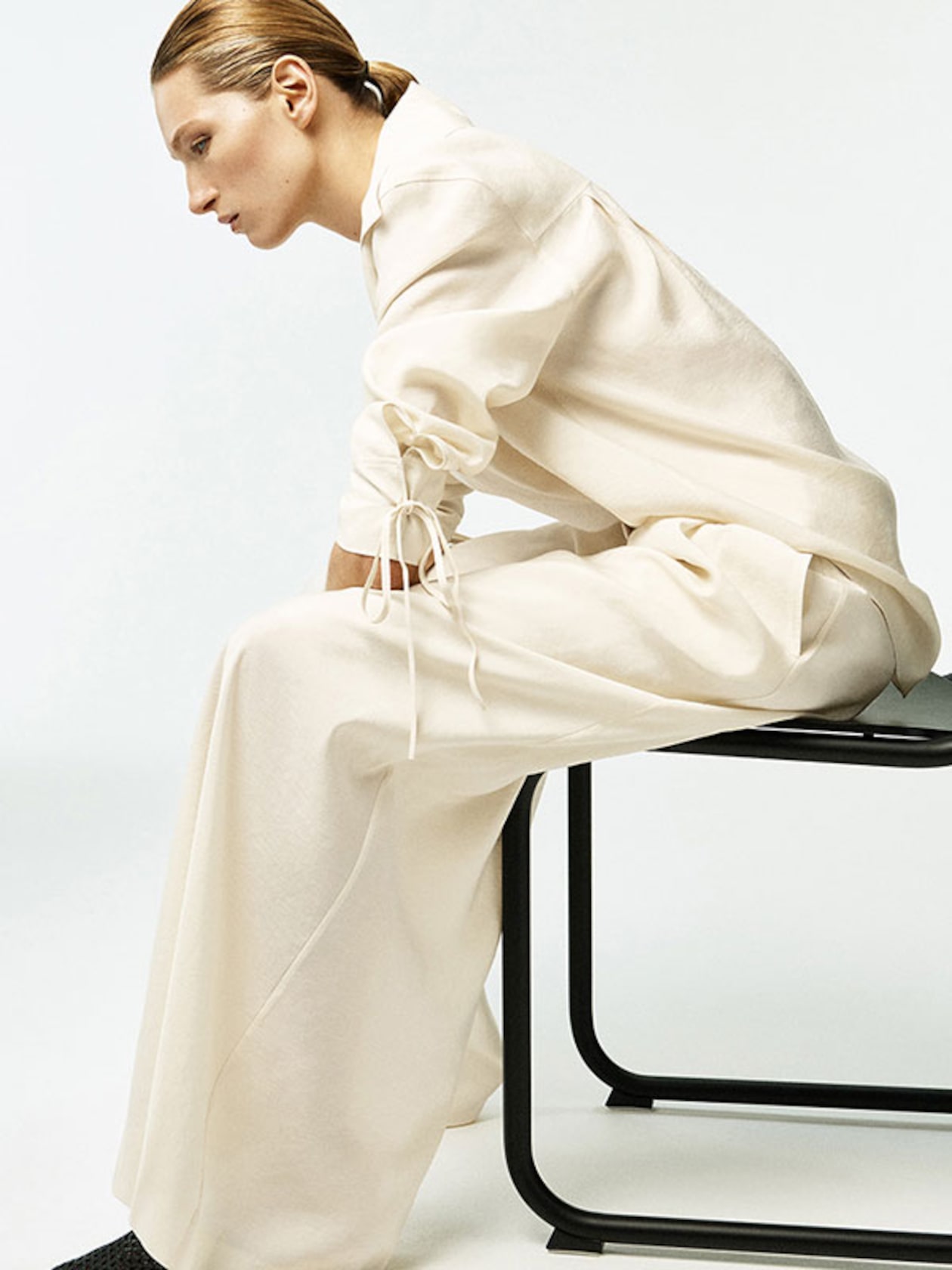 Model in a loose, light-colored outfit sits on a black chair, leaning forward against a plain backdrop.
