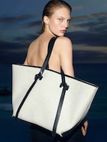 Placeholder for: A person with slicked-back hair poses with a large beige tote bag featuring black straps, set against a twilight ocean backdrop.