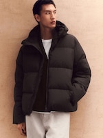 Placeholder for: Person in a black puffer jacket and white pants stands against a beige wall, looking off to the side.