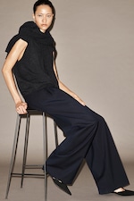 Placeholder for: Woman in dark sleeveless top and wide-leg pants sits on a high stool against a neutral background, with hair tied back.