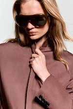 Placeholder for: Woman wearing large sunglasses and a brown jacket, with long blonde hair cascading over her shoulders, holding her jacket collar.