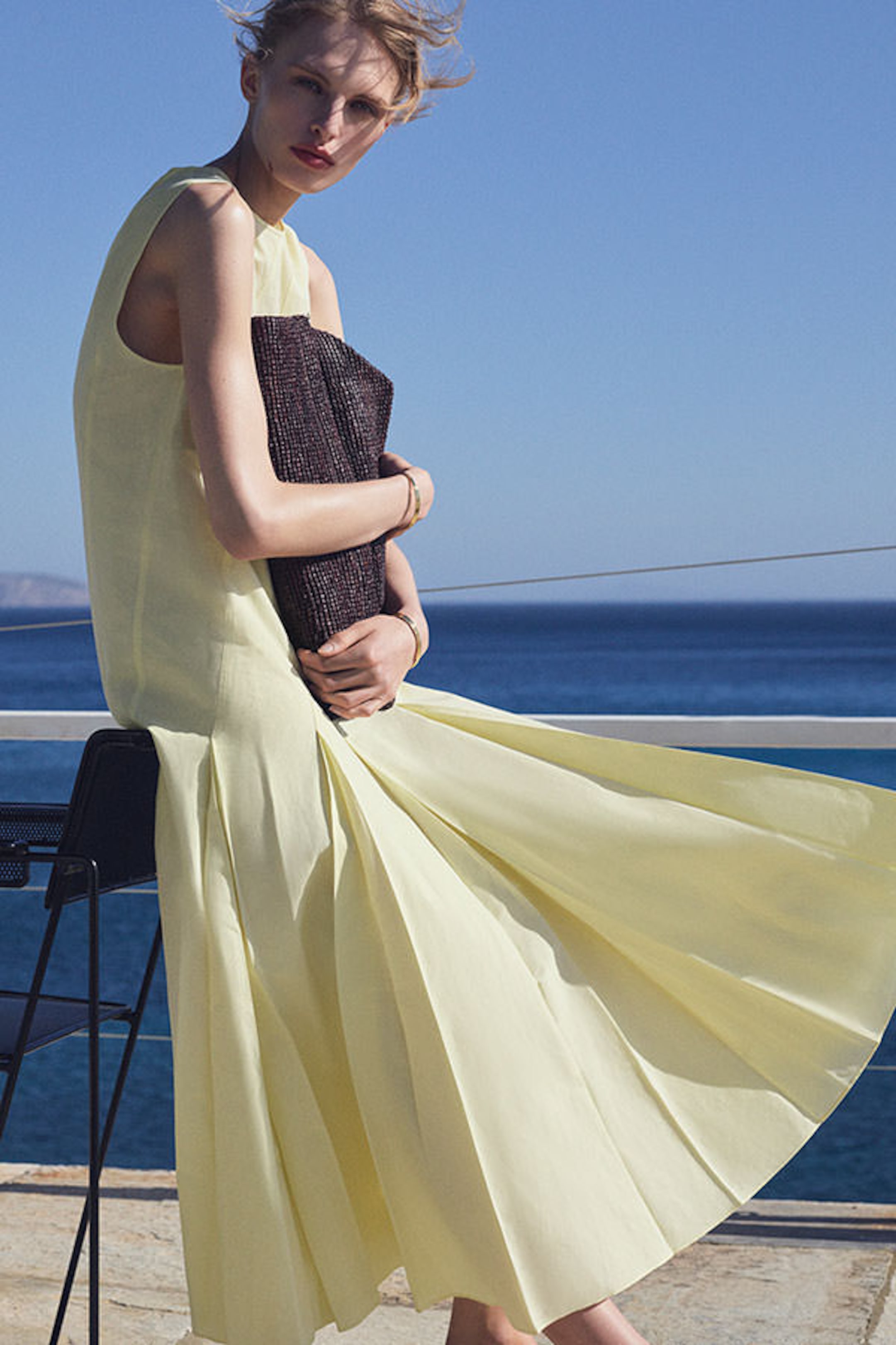 A woman in a flowing yellow dress sits near the ocean, holding a dark clutch, with wind blowing her hair and the hem of her dress.