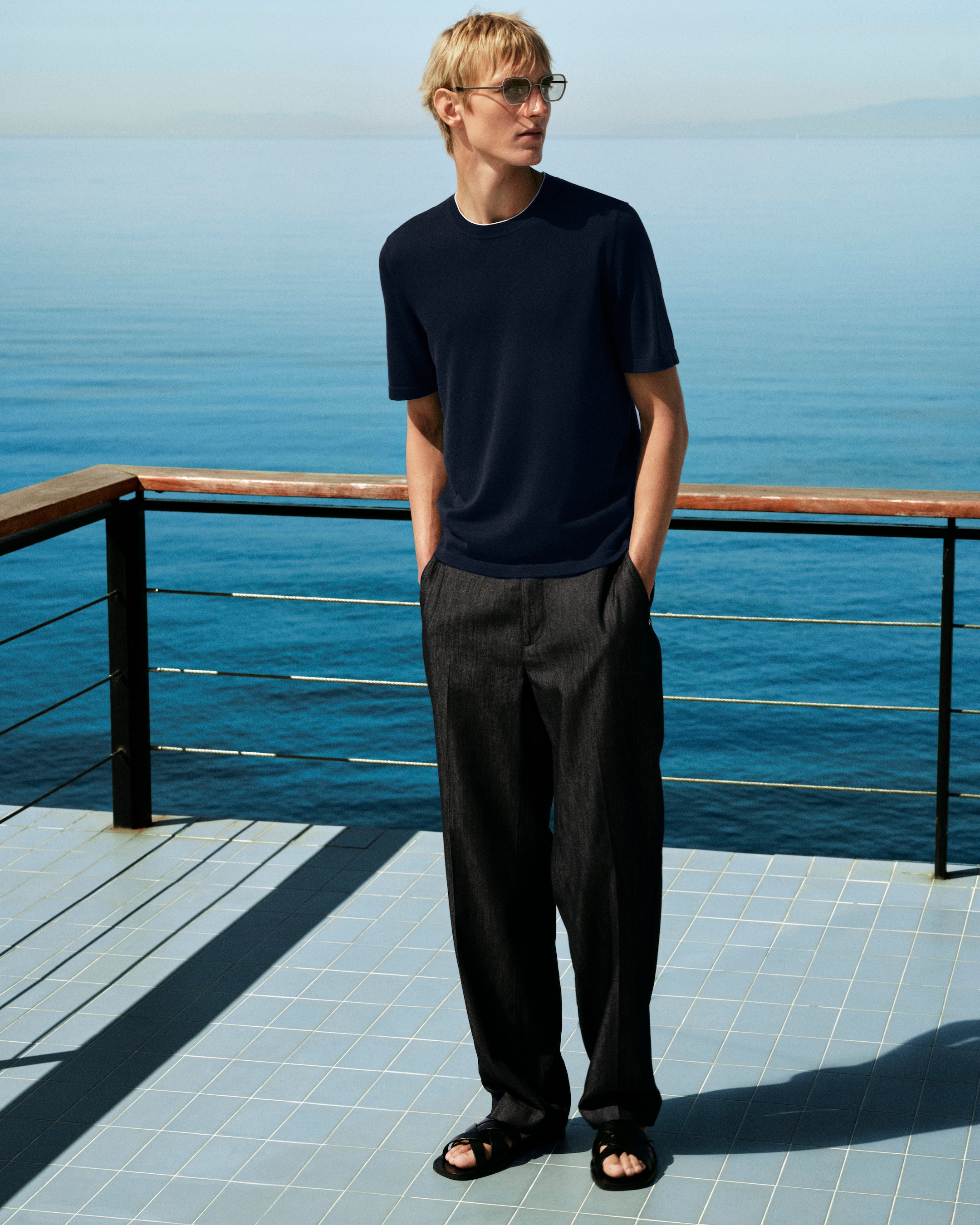 A person in a navy T-shirt and dark pants stands on a tiled balcony, overlooking a calm blue sea under clear skies.