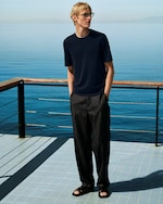 Placeholder for: A person in a navy T-shirt and dark pants stands on a tiled balcony, overlooking a calm blue sea under clear skies.